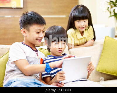 three asian children two little boy and one little girl sitting on couch at home playing video game with digital tablet, focus on the little boyin the Stock Photo
