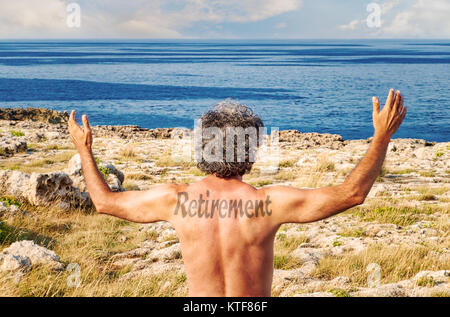 Retirement word tatoo on back of mature man raising hands on seaside while looking to the horizon Stock Photo