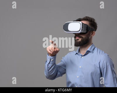 Business man wearing virtual reality glasses points finger and interact with augmented reality touch screen with copyspace over gray background. Stock Photo