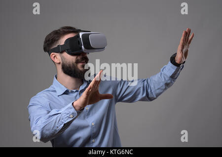 Amazed young adult bearded smart casual man wearing virtual reality vr headset gesturing hands and looking at copyspace over gray background. Stock Photo