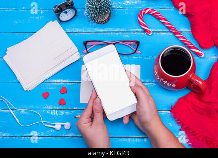 Two female hands hold a white smartphone with a blank screen on a blue wooden table, next is a red mug with coffee and empty vintage postcards Stock Photo