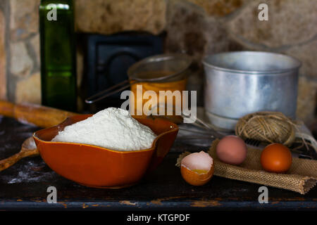 White wheat flour in ceramic ware, broken egg with the yolk, whole eggs and cooking utensils for cooking dough on a background of a stone oven. Rustic Stock Photo