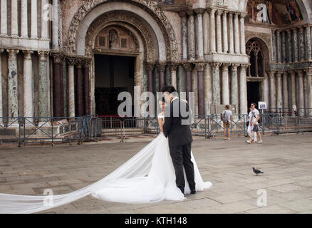 Newly married Asian couple with their wedding outfits posing for a photographer in front of Saint Mark's Basilica (Basilica di San Marco) in Venice. Stock Photo