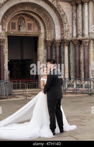 Newly married Asian couple with their wedding outfits posing for a photographer in front of Saint Mark's Basilica (Basilica di San Marco) in Venice. Stock Photo