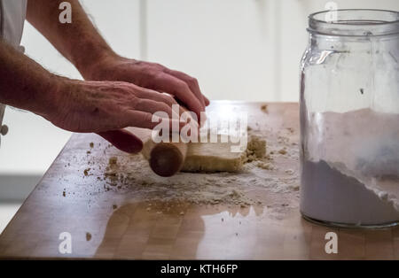 Close up of older in chef whites rolling dough with a rolling pin making pastry on a wooden block with a flour jar in a kitchen Stock Photo