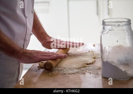 Close up of older in chef whites rolling dough with a rolling pin making pastry on a wooden block with a flour jar in a kitchen Stock Photo