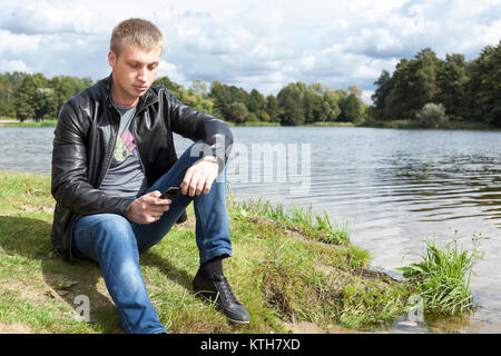 Blond Caucasian man in black leather jacket sitting on river bank with mobile phone in hands Stock Photo