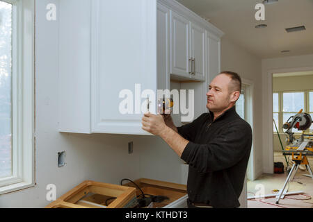 The worker sets a new handle on the white cabinet with a screwdriver installing kitchen cabinets Stock Photo