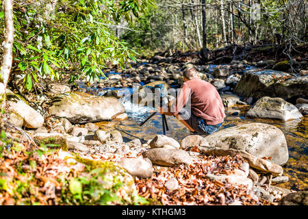 Young man nature photographer with camera and tripod setting up by shallow rock stream with running water and waterfall in autumn with foliage, stones Stock Photo