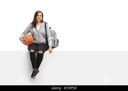 Teenage girl with a basketball and a backpack seated on a panel isolated on white background