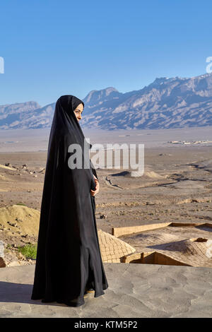 Yazd, Iran - April 21, 2017: One unknown Iranian Muslim girl, dressed in religious veil chador, stands on roof of  clay rural house, against backdrop  Stock Photo