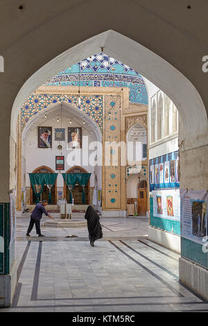 Yazd, Iran - April 22, 2017:  A Muslim woman in a black Islamic veil passes by a cleaner who wipes the marble floor at the entrance to the mosque. Stock Photo