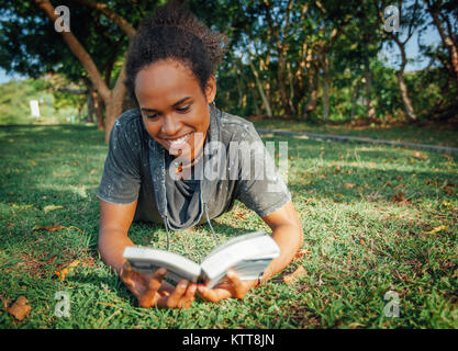 Beautiful young woman with afro hairstyle and book in her hands lying on the grass in the park on sunny summer day Stock Photo