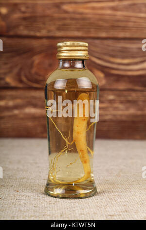Close up of one glass jar bottle of ginseng root essence extraction liquor with golden cap, on canvas tablecloth over brown wooden background, low ang