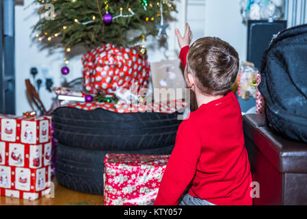 Boy in red christmas sweater reaching to gifts under Christmas tree. Stock Photo