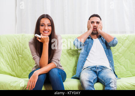 Young couple is sitting on sofa at home. Man is frustrated because he does not like what his woman is watching on TV. Stock Photo