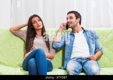 Young couple is sitting on sofa at home. Woman is getting bored while man is typing on phone. Stock Photo
