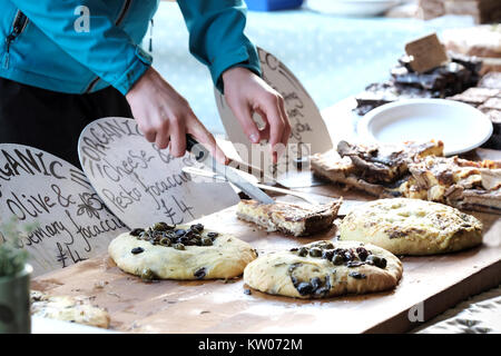 A man working on an open air stall selling Organic Focaccia flatbread. He's using a knife to cut a portion for a customer. The breads are all priced Stock Photo