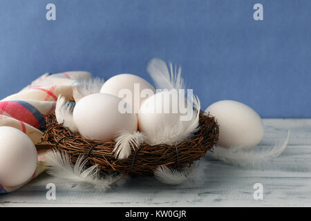 White chicken eggs in the nest with feathers on white wooden table over purple background. Copy space Stock Photo
