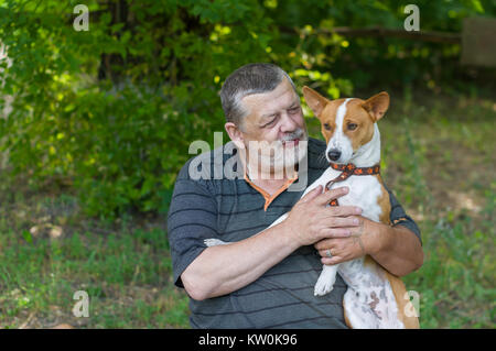 Bearded senior man talking to his cute dog (basenji) taking it in the hands while resting in summer park