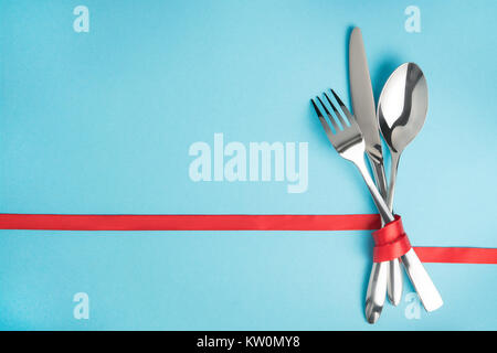 Fork, spoon and knife tied with a red ribbon on blue background with copy space. Food, restaurant and table setting theme Stock Photo