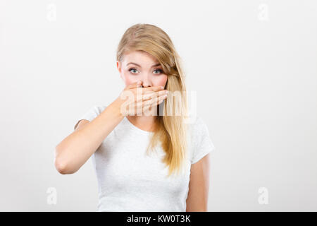 Saying bad things, shocking news concept. Ashamed young blonde woman having hand on her mouth. Studio shot on grey background. Stock Photo