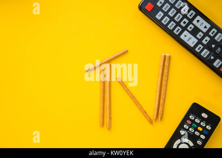 Weekend, leisure and hobby concept. A TV inscription made of bread sticks and remote controls on a bright one-colore yellow background. Space for your Stock Photo