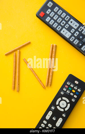 Weekend, leisure and hobby concept. A TV inscription made of bread sticks and remote controls on a bright one-colore yellow background. Vertical orien Stock Photo