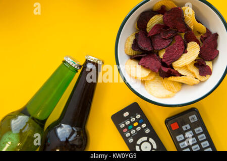 watching tv shows. Weekend, hobby and leisure concept. A bowl of potato and beetroot chips, two bottles of beer and two remote controls on a bright ye Stock Photo