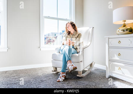 Bright white modern rocking chair in nursery room with chest of drawers, decorations in model staging home, apartment or house, one young woman sittin Stock Photo