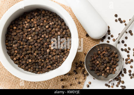 Raw, natural, unprocessed black pepper peppercorns in mortar and metal spoon on white wooden table Stock Photo