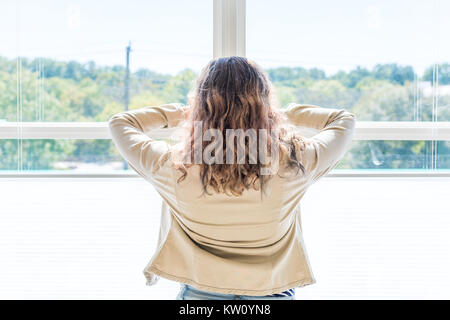 Back of one young woman standing in white, bright room of house, home, apartment looking through the sunny large window with blinds Stock Photo