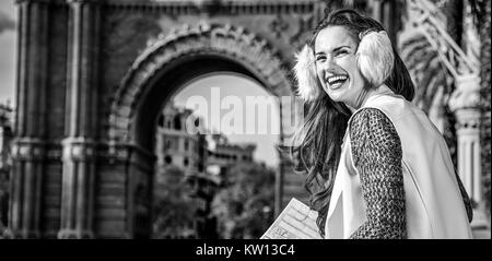 in Barcelona for a perfect winter. smiling elegant woman in earmuffs near Arc de Triomf in Barcelona, Spain with map looking into the distance Stock Photo