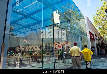 On a sunny day, two men--one wearing a Stanford University baseball cap--walk past the entrance to the flagship Apple electronics store on University Avenue in downtown Palo Alto, California, 2016. Shoppers are visible browsing inside the store. Stock Photo