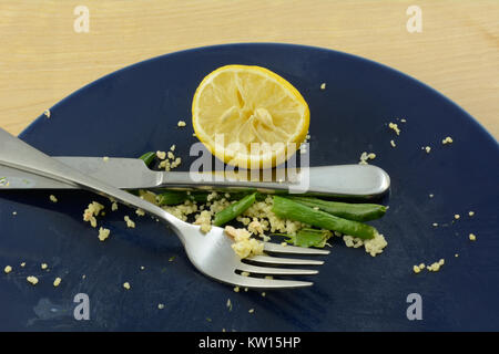 Last uneaten food of green beans,couscous, squeezed lemon half with knife and fork placed down on blue plate Stock Photo