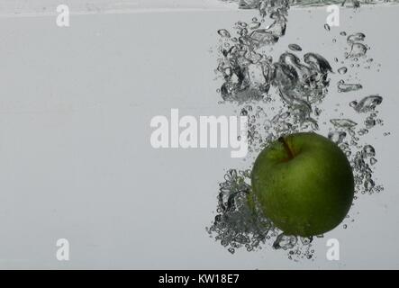 Green apple falling into water at an angle, surrounded by air bubbles. Light grey background Stock Photo