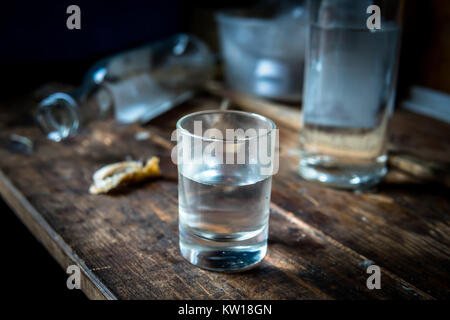 A small stack of vodka close up on the old dirty wooden table amid empty bottles lying on the table. Alcoholism concept. Stock Photo