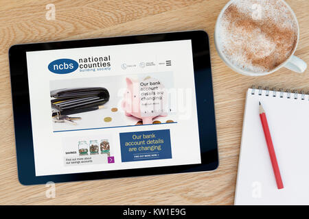 The NCBS (National Counties Building Society) website on an iPad, resting on a wooden table beside a notepad pencil and cup of coffee (Editorial only) Stock Photo