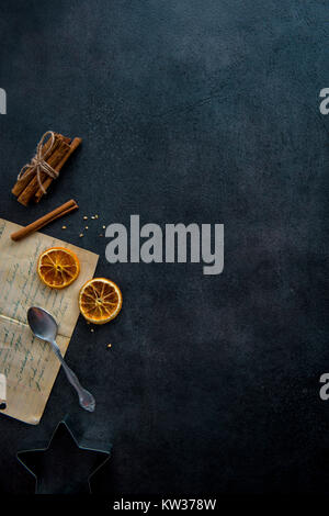 Food preparation. Old recipe, teaspoon, cookie cutter and some ingredients (cinnamon sticks, dried orange slices) on dark modern table from above. Stock Photo