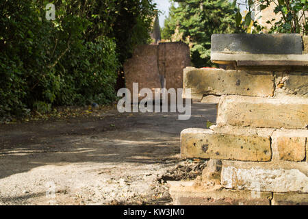 A close up of a broken brick wall with a boarded up house in the background, shadows casted on the ground. Trees surrounding pathway. Creepy suburban. Stock Photo