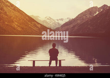 man sitting alone on park bench under tree. backpacker resting on the shore of the lake enjoying the view. The concept of loneliness. Stock Photo