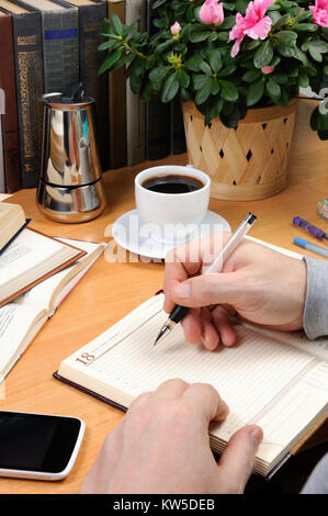 a man holds a pen and takes notes in a notebook on a desk in his office, surrounded by books, notebooks with a cup of black coffee Stock Photo