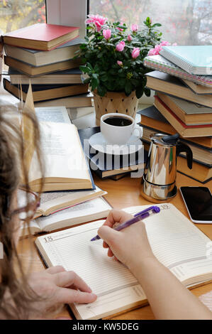 A woman holds a pen and takes notes in a notebook on a desk in her office, surrounded by books, notebooks with a cup of black coffee Stock Photo