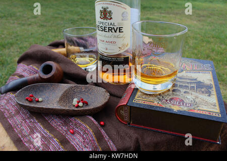 Still life - two glasses with bottle of whisky, wooden pipe and bowl and a book arranged on a garden table Stock Photo