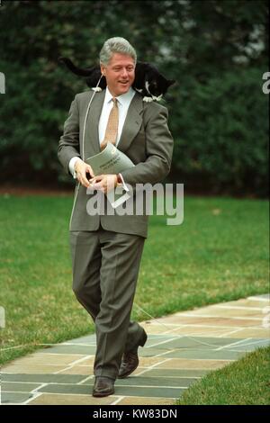 A smiling President William Jefferson Clinton wearing taupe suit walks down a stone pathway on the White House grounds with Socks the Cat, with black fur, white face, and long string leash dangling, perched upon his shoulders, holding a packet labeled 'Presidential Statement, ' Washington, District of Columbia, March 7, 1995. Stock Photo