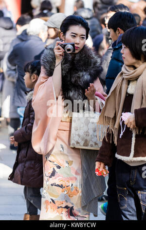 Japan, Nishinomiya Shinto shrine. New year's Day, Shogatsu. Young woman in kimono and black fur collar standing in the crowd holding point and shoot camera. Daytime. Stock Photo