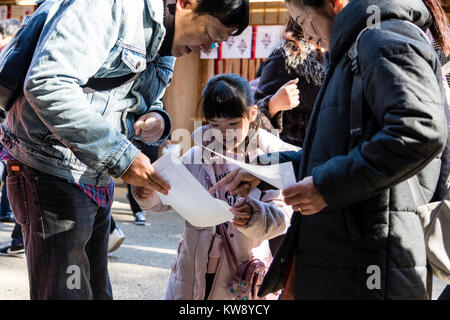 Japan, Nishinomiya Shinto Shrine. New year's day, Shogatsu. Family on Hatsumode visit, (first visit of the new year), reading a fortune paper, Omikuji to their child daughter. She looks happy and smiling as they bend to explain. Stock Photo