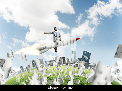 Conceptual image of young businessman in suit flying on rocket among flying papers with cityscape and blue sky on background. Stock Photo