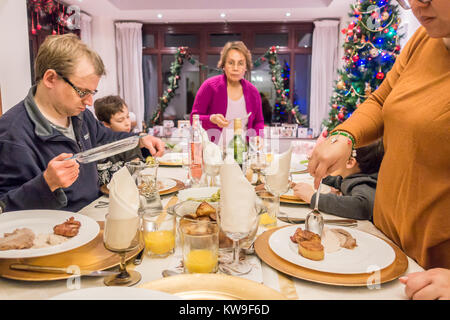 A mixed race family serving out Christmas dinner on a dining table. Plates sit on gold coloured chargers Glasses an napkins are on the table. Stock Photo
