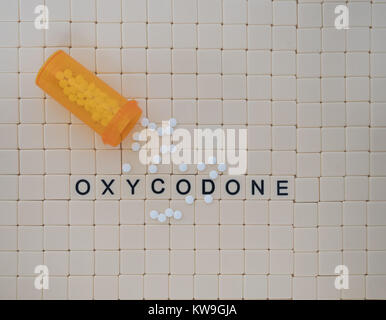 Oxycodone spelled in capital letters in beige tiles with black letters.  An open bottle of tablets with scattered pills is photographed from above. Stock Photo